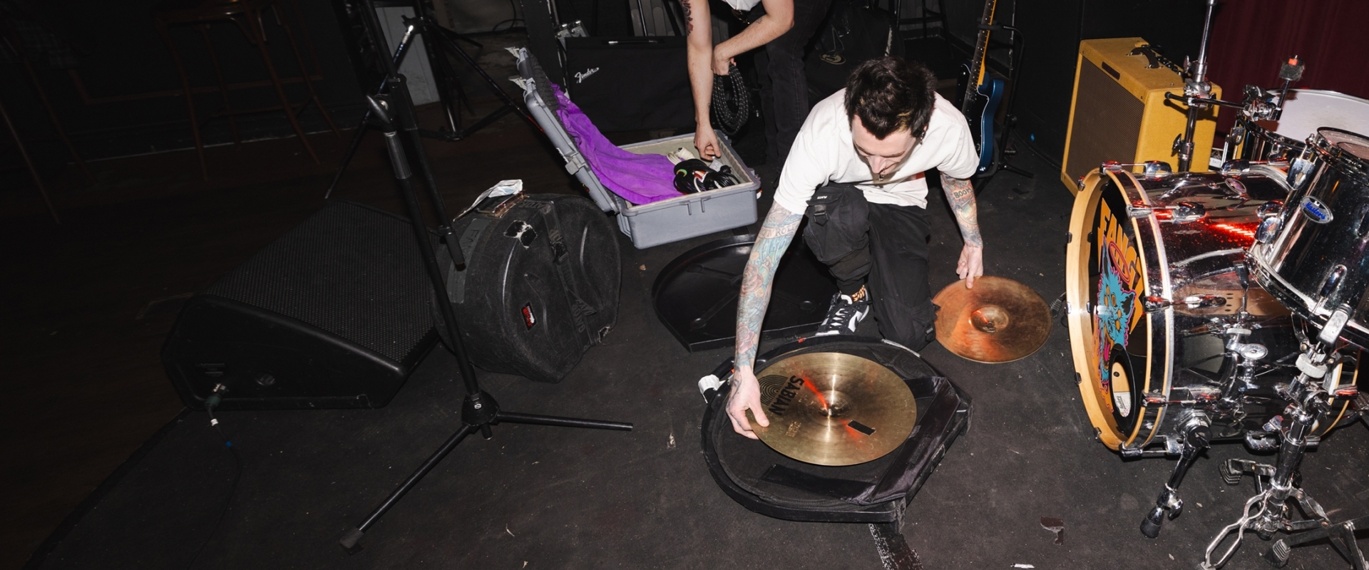 Band member kneeling down to set up or pack away gold drum cymbals and music equipment on a stage.