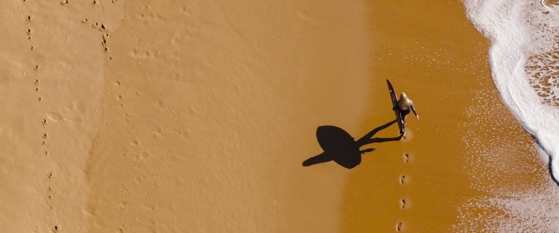 Aerial view of a person with a surfboard casting a long shadow, walking along the wet sand next to the ocean waves.